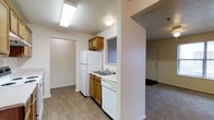 This single-floor space offers a practical and efficient layout designed to maximize functionality. The kitchen serves as a central feature, equipped with warm honey-toned wood cabinetry, a white refrigerator, a dishwasher, and a double-basin sink set beneath a window that allows natural light to filter in. A textured ceiling with overhead fluorescent lighting ensures the kitchen remains bright and well-lit. The hardwood-style flooring in the kitchen adds a clean, modern touch and transitions smoothly into adjacent areas. A partial wall divides the kitchen from the carpeted living space, which features a front entry door and closet, providing a defined yet open sense of flow throughout the unit. The neutral white and beige color palette throughout the space creates a calm, cohesive ambiance that lends itself well to personalization. A sliding closet door is visible near the kitchen, offering convenient storage. The overall design is straightforward and functional, with each area thoughtfully connected to support comfortable everyday living on a single level.
