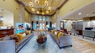 This impressive single-story community clubhouse showcases a thoughtful blend of rustic and contemporary design throughout its well-appointed spaces. The centerpiece is a grand lounge area with dramatically soaring ceilings, striking natural stone columns, and an eye-catching multi-arm chandelier that anchors the room. Plush gray sofas accented with gold and neutral throw pillows are arranged around a decorative round coffee table atop a large area rug, creating an inviting gathering space. Tall windows and glass doors flood the interior with natural light, while warm hardwood flooring flows throughout. A cozy dining nook features blue upholstered chairs around a round wood table, framed by exposed stone walls and elegant drapery. The full kitchen is outfitted with rich wood cabinetry, dark granite countertops, a central island with pendant lighting, and modern appliances. A dedicated business center provides multiple workstations with built-in wood cabinetry and polished concrete floors. The fitness center is well-equipped with cardio machines, free weights, and wall-mounted TVs. Outside, a spacious stone-paved patio with comfortable seating offers a relaxing retreat surrounded by the building's handsome natural stone façade.