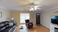 This single-story, 3-bedroom, 3-bathroom home offers a warm and functional layout designed for comfortable everyday living. The living room sets an inviting tone with its rich hardwood floors, crown molding, and a ceiling fan with light kit that keeps the space bright and well-ventilated. A large window fitted with blinds allows natural light to filter in, creating a cheerful atmosphere throughout the day. The entryway features a classic dark front door with a decorative glass inset, adding a touch of character to the home's interior. The living area comfortably accommodates seating, including a plush leather recliner and sofa, centered around a wooden coffee table — ideal for relaxing or entertaining guests. The neutral wall tones complement the warm wood flooring, giving the home a cohesive and timeless aesthetic. With three full bathrooms to accompany the three bedrooms, this home provides ample space and convenience for families or those who appreciate having room to spread out. The single-floor layout ensures easy accessibility throughout, making it a practical and comfortable place to call home.