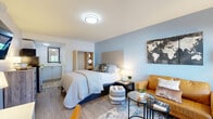 This well-appointed studio suite makes smart use of its compact footprint, offering a thoughtfully organized layout that includes a bedroom, kitchenette, and full bathroom all on a single floor. The main living area is anchored by a full-sized bed with a warm wood-toned headboard and an open wardrobe unit with integrated shelving, set against soothing light blue accent walls and light wood-grain flooring. A cozy sitting area shares the same open space, featuring a caramel leather loveseat, a glass-top coffee table, and a decorative world map art piece, creating a relaxed yet functional living environment. Natural light filters in through a window offering a pleasant outdoor view, adding an airy feel to the space. The kitchenette is clean and efficient, outfitted with white cabinetry, a dual-burner induction cooktop, an L-shaped countertop with a stainless steel sink, and ample upper cabinet storage. The bathroom is crisp and modern, featuring white subway tile surrounding a full bathtub and shower combo, wood-look tile flooring, and a vanity with storage. Throughout the suite, the design is cohesive, combining neutral tones, modern fixtures, and practical storage solutions to maximize comfort within a compact living space.