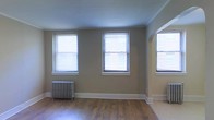 This single-floor space showcases a blend of classic and functional design. The main living area is bathed in natural light, courtesy of multiple large windows fitted with horizontal blinds that allow for adjustable brightness throughout the day. The warm-toned hardwood flooring runs throughout, adding a timeless character to the space.

A notable architectural feature is the elegant arched entryway, which adds a touch of classic charm and creates a seamless visual flow between areas. The neutral, earthy wall tones complement the flooring and contribute to a calm, welcoming atmosphere.

A traditional cast-iron radiator sits along one wall, serving as both a functional heating element and a distinctive period detail that speaks to the building's heritage. Clean white baseboard trim outlines the perimeter of the room, providing a crisp contrast to the warmer tones throughout.

The ceilings are smooth and generous in height, further enhancing the open and airy feel of the space. Overall, the design strikes a balance between historic architectural character and practical living, making for a space that is both full of personality and comfortably livable.