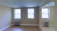 This single-floor space showcases a blend of classic and functional design. The main living area is bathed in natural light, courtesy of multiple large windows fitted with horizontal blinds that allow for adjustable brightness throughout the day. The warm-toned hardwood flooring runs throughout, adding a timeless character to the space.

A notable architectural feature is the elegant arched entryway, which adds a touch of classic charm and creates a seamless visual flow between areas. The neutral, earthy wall tones complement the flooring and contribute to a calm, welcoming atmosphere.

A traditional cast-iron radiator sits along one wall, serving as both a functional heating element and a distinctive period detail that speaks to the building's heritage. Clean white baseboard trim outlines the perimeter of the room, providing a crisp contrast to the warmer tones throughout.

The ceilings are smooth and generous in height, further enhancing the open and airy feel of the space. Overall, the design strikes a balance between historic architectural character and practical living, making for a space that is both full of personality and comfortably livable.