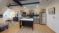 Located at 201 S Huntington Ave in Boston's Jamaica Plain neighborhood, this single-floor unit blends industrial character with modern finishes throughout. The open living area is defined by striking exposed steel beams and a bold black structural column, lending a loft-like atmosphere to the space. Light hardwood flooring runs continuously throughout, complementing the bright white walls and creating a warm, cohesive feel.

The kitchen is thoughtfully appointed with gray shaker-style cabinetry, quartz countertops, a deep navy mosaic tile backsplash, and a full suite of stainless steel appliances. A mobile island with a quartz top provides additional prep and counter space. Recessed lighting above highlights the exposed beam ceiling structure overhead.

The bedroom features distinctive angled roofline ceilings with exposed dark beams, adding architectural interest and a cozy, tucked-away character. The living area benefits from multiple windows that bring in natural light and offer views of surrounding greenery.

The bathroom is finished with hexagonal tile flooring, a warm wood vanity, matte black hardware accents, and a glass-enclosed walk-in shower clad in classic white subway tile. A dark painted ceiling adds a modern, contrasting design touch. The combination of raw industrial elements and refined modern updates gives this space a distinctive and dynamic character.
