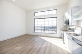 This single-floor space offers a thoughtfully designed open layout finished with light hardwood flooring and crisp white walls throughout, creating a bright and cohesive aesthetic. The kitchen is a standout feature, equipped with stainless steel appliances including a range, microwave, dishwasher, and refrigerator, complemented by white shaker-style cabinetry, a subway tile backsplash, and a gold-toned faucet at the sink. A dramatic floor-to-ceiling industrial-style window floods the kitchen and living area with abundant natural light while framing views of the surrounding urban landscape. A sliding barn door adds a modern, space-efficient architectural detail to the hallway, which also includes a built-in countertop nook. The open living area is generously proportioned with soaring ceilings and a ceiling fan overhead. The bathroom features white shaker cabinetry with matte black hardware, a single vanity with a neutral countertop, a large mirror, and a walk-in shower with brushed nickel fixtures. Additional rooms present clean, minimalist spaces with angled ceiling lines and ample room for flexible use. Overall, the space delivers a contemporary, light-filled atmosphere with modern finishes.