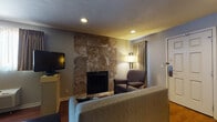 Located at 711 South Main Street in Wichita, Kansas, this single-floor guest suite offers a thoughtfully designed layout that combines comfort and functionality. The living area serves as a welcoming focal point, anchored by a striking floor-to-ceiling stone-tiled fireplace that adds warmth and character to the space. A sofa, upholstered armchair, and flat-screen TV complete this relaxing area, all set atop rich hardwood-style flooring that flows throughout the suite. The sleeping area features a generously sized bed set against a bold accent wall, complemented by a wall-mounted TV, a window with curtains allowing natural light, and an in-unit climate control unit for personalized comfort. A full kitchen provides practical amenities including dark wood cabinetry, a tile backsplash, full-sized refrigerator, electric range, dishwasher, and a sink with ample counter space — ideal for extended stays. The bathroom is clean and functional, offering a shower/tub combination with coordinated linens and built-in shelving. Recessed lighting and neutral grey walls throughout give the suite a consistent, understated aesthetic, while the open-concept arrangement of the living and sleeping zones creates a sense of spaciousness well-suited for both short and longer-term visits.