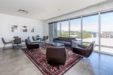 Located at 219 E Grand Ave in downtown Des Moines, Iowa, this modern single-floor living space offers a thoughtfully designed interior with a clean, contemporary aesthetic. The open layout is bathed in natural light, thanks to expansive floor-to-ceiling glass walls that frame panoramic views of the Des Moines skyline and surrounding cityscape.

The living room is divided into two distinct functional zones. The lounge area features leather seating arranged around a glass-top coffee table, anchored by a vibrant patterned area rug. A wall-mounted flat-screen TV sits above a light wood media console, while a curated gallery of framed artwork lines the crisp white walls, adding a refined artistic touch. The second zone serves as a recreation and social space, centered around a full-size billiards table set atop a geometric patterned rug, with additional seating nearby.

Polished concrete floors run throughout, complementing the minimalist white walls and recessed linear lighting. Vaulted ceiling details in one section of the space add architectural interest and a sense of openness. Glass sliding doors provide direct access to an outdoor terrace, seamlessly connecting the interior to the exterior. The overall ambiance is sophisticated, bright, and versatile — well-suited for both social gatherings and relaxed everyday use.
