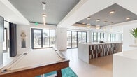 Located at 9324 South Breakwater Boulevard in Oak Creek, Wisconsin, this contemporary community clubhouse offers a thoughtfully designed single-floor layout that blends entertainment, relaxation, and social gathering seamlessly. The open-plan space is anchored by a stylish lounge area featuring light hardwood flooring, a striking dark accent ceiling, a wall-mounted fireplace flanked by vertical shiplap paneling, and a wall-mounted television — all illuminated by a sculptural brass chandelier. A full-size pool table atop a vibrant teal area rug serves as a central focal point in the game zone, while a dedicated shuffleboard room with floor-to-ceiling windows and a bar-height counter with seating provides additional recreational options. A colorfully tiled bar-style counter adds a bold design element near the dining area, which is bathed in natural light from expansive black-framed sliding glass doors that open directly onto an outdoor terrace with patio seating. The white walls and light-toned flooring throughout create a bright, airy ambiance, while the carefully placed pendant lighting and recessed fixtures add warmth and depth. The result is a well-appointed, versatile gathering space ideal for both casual socializing and organized events.