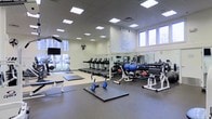 This fitness center offers a comprehensive and well-organized workout environment spread across a single floor. The space is bright and airy, benefiting from an abundance of natural light streaming through large windows that line the walls, complemented by a grid of recessed ceiling panel lights that ensure consistent illumination throughout.

The cardio zone features a row of treadmills aligned along one wall, providing users with a dedicated area for aerobic exercise. Adjacent to this, a large wall-to-wall mirror expands the sense of space and allows for form monitoring during workouts. The free weights section is well-stocked with a full dumbbell rack, medicine balls, and dedicated weight benches, catering to a wide range of strength training needs.

Functional training mats are laid out on the open floor area, offering ample room for stretching, bodyweight exercises, or dynamic movements. A mounted flat-screen television keeps members informed or entertained during their sessions. A standing fan adds to the ventilation and comfort of the space. The flooring transitions between rubberized and smooth surfaces to suit different workout activities. Gym rules are prominently posted on the wall, reflecting a well-managed and community-oriented facility. The overall ambiance is clean, practical, and welcoming.