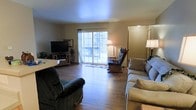 This single-floor space centers around a generously sized living room that immediately conveys a sense of openness and comfort. The room features rich hardwood flooring that extends throughout, lending warmth and a classic aesthetic to the interior. Neutral-toned walls complement the natural light that pours in through a large sliding glass door, which serves as a prominent architectural feature and provides a direct view of the outdoor surroundings. The sliding door also offers convenient access to what appears to be an exterior deck or patio area, enhancing the indoor-outdoor connection. The living room is well-suited for both relaxation and entertaining, with ample space to accommodate multiple seating arrangements. Floor lamps positioned around the room provide warm ambient lighting, adding to the inviting atmosphere. A dedicated area near the sliding door accommodates a media console and television, creating a functional entertainment zone. The layout flows naturally, allowing for clear sightlines across the room. Overall, the space presents a welcoming and practical environment, characterized by its warm tones, natural light, durable flooring, and thoughtful layout that balances comfort with everyday functionality.