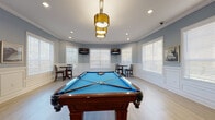 Located at 10800 Torino Dr in Trinity, FL, this single-floor recreational game room is thoughtfully designed to serve as a premier gathering and entertainment space. The room's most prominent feature is a full-size billiards table with a vivid blue felt surface, positioned at the heart of the space beneath a striking gold pendant light fixture complemented by recessed lighting throughout. Light hardwood flooring runs across the entire room, creating a clean, contemporary foundation that contrasts elegantly with the soft blue-gray walls. Detailed white wainscoting panels line the lower half of the walls, adding a refined architectural character to the space. Multiple wall-mounted flat-screen televisions are strategically placed throughout the room, ensuring visibility from various seating areas. Cozy bistro-style tables paired with counter-height and standard chairs are arranged in the angled bay corners of the room, offering comfortable spots for conversation or viewing. Large windows fitted with white plantation-style blinds allow abundant natural light to fill the space while maintaining privacy. A dark wood storage cabinet provides practical storage for pool cues and accessories. Double glass-panel French doors serve as the main entry, offering an open, welcoming transition into the room. Crown molding details complete the polished, community-oriented ambiance of this well-appointed game room.