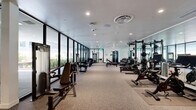 Located at 1650 Lincoln Blvd in Santa Monica, California, this single-floor fitness facility offers a generously sized, comprehensively equipped exercise space designed to support a wide range of training styles. The layout is thoughtfully divided into distinct zones, each tailored for specific workout disciplines.

The primary gym area features carpet flooring and is lined with floor-to-ceiling mirrored walls that amplify the sense of space. This section is equipped with a full array of cardio machines — including treadmills, ellipticals, stair climbers, and cycling equipment — alongside a robust selection of strength training machines, free weights, and adjustable benches. Recessed lighting ensures a bright, well-lit environment throughout.

A dedicated turf zone with artificial grass flooring provides space for functional and high-intensity training, housing equipment such as rowing machines, cable trainers, sled pushers, and plyo boxes. This area transitions into a semi-outdoor corridor featuring large planters with tall ornamental grasses and open views to an adjacent courtyard.

The courtyard area is characterized by its clean, modern architectural lines, smooth concrete ceiling, large-format tile flooring, and abundant natural light filtering in from the open surroundings. Greenery throughout softens the contemporary aesthetic, creating an inviting atmosphere that bridges indoor training with the outdoor Santa Monica environment.