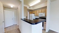 This single-floor space centers around a well-organized kitchen that serves as the focal point of the layout. The kitchen features a wraparound peninsula with a dark countertop, providing ample workspace and a natural division between the kitchen and the adjacent living area. Light wood cabinetry lines the walls, offering generous storage, while white appliances — including a refrigerator, microwave, and range — contribute to a clean, cohesive look. Overhead fluorescent lighting ensures the kitchen remains well-lit and practical. The layout is designed for efficiency, with the appliances and cabinetry arranged to maximize functionality. Neutral-toned walls and light tile flooring extend throughout the space, creating a consistent and understated aesthetic. A hallway visible from the kitchen leads to additional areas of the unit, hinting at a straightforward, accessible floor plan. The overall ambiance is simple and utilitarian, emphasizing practicality while maintaining a tidy, open feel suitable for everyday living.