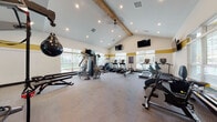 This spacious fitness center is designed to support a full range of workout routines in a bright, inviting environment. The room's most striking architectural feature is its vaulted ceiling accented by a prominent exposed wood beam, which adds warmth and character to the space. Recessed lighting and a ceiling fan complement the abundant natural light that streams in through large windows lining multiple walls, offering views of the surrounding outdoor greenery.

The interior is finished with neutral carpet flooring and walls adorned with a distinctive gold accent stripe, giving the space a cohesive and energetic aesthetic. Multiple wall-mounted flat-screen televisions are positioned throughout, allowing users to stay entertained during their workouts.

The cardio zone is well-stocked with treadmills, elliptical machines, stair climbers, rowing machines, and stationary bikes, including True-brand equipment. The strength training area features a multi-station cable machine, adjustable benches, a dumbbell rack with a full range of free weights, and a speed bag for boxing training. Full-length mirrors along one wall enhance the sense of space and allow users to monitor their form. A posted fitness center rules board reinforces the facility's organized, community-oriented atmosphere.