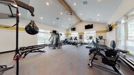 This spacious fitness center is designed to support a full range of workout routines in a bright, inviting environment. The room's most striking architectural feature is its vaulted ceiling accented by a prominent exposed wood beam, which adds warmth and character to the space. Recessed lighting and a ceiling fan complement the abundant natural light that streams in through large windows lining multiple walls, offering views of the surrounding outdoor greenery.

The interior is finished with neutral carpet flooring and walls adorned with a distinctive gold accent stripe, giving the space a cohesive and energetic aesthetic. Multiple wall-mounted flat-screen televisions are positioned throughout, allowing users to stay entertained during their workouts.

The cardio zone is well-stocked with treadmills, elliptical machines, stair climbers, rowing machines, and stationary bikes, including True-brand equipment. The strength training area features a multi-station cable machine, adjustable benches, a dumbbell rack with a full range of free weights, and a speed bag for boxing training. Full-length mirrors along one wall enhance the sense of space and allow users to monitor their form. A posted fitness center rules board reinforces the facility's organized, community-oriented atmosphere.