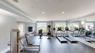 This fitness center offers a comprehensive workout environment thoughtfully designed to support a wide range of exercise routines. The space is finished with soft blue-gray walls, white crown molding, and recessed lighting that creates a bright, energizing atmosphere. Durable rubber flooring spans the entire area, providing both safety and comfort underfoot.

The cardio zone is lined with multiple treadmills, elliptical trainers, and a stationary bike, all positioned facing large double-hung windows that frame views of lush greenery outdoors — allowing natural light to fill the room during workouts. A wall clock and wall-mounted televisions add functional convenience throughout the space.

The strength training section is equally well-appointed, featuring a cable crossover machine, a variety of selectorized weight machines targeting different muscle groups, a rowing machine, free weights with a dedicated dumbbell rack, and flat benches for versatile use. A full-length wall mirror in the strength area enhances both the sense of space and allows for proper form checks during exercise.

Wall sconces supplement the recessed lighting in the strength zone, contributing to an inviting ambiance. The facility is neatly organized, clean, and spacious, offering users ample room to move between stations comfortably. Overall, this fitness center delivers a professional and fully functional environment for a complete workout experience.
