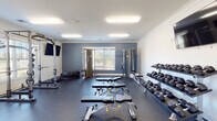 Located at 134 Turning Leaf Way in Sanford, NC, this single-floor fitness center is thoughtfully designed to accommodate a wide range of workout needs. The facility is divided into distinct functional zones, each finished with attention to practicality and comfort.

The primary strength training area features durable rubber flooring in a deep charcoal tone, complemented by blue-gray accent walls and crown molding. This zone is equipped with a full power rack loaded with weight plates, a comprehensive dumbbell rack spanning a wide weight range, multiple adjustable benches, and a cable machine. Wall-mounted flat-screen TVs provide entertainment during workouts, while multiple windows allow natural light to filter in and offer views of the surrounding neighborhood.

A separate cardio and flex area transitions to light-toned carpet tile flooring, maintaining a clean, bright aesthetic with crisp white walls. This section houses a rowing machine, a stair climber, and exercise balls, providing options for low-impact and functional training. A wall-mounted TV and a dedicated water fountain station serve this area as well.

A glass-paneled entry door provides access directly from outside, and posted fitness room rules ensure an orderly environment. The overall facility presents a well-organized, community-oriented fitness space suitable for a variety of fitness routines.