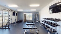 Located at 134 Turning Leaf Way in Sanford, NC, this single-floor fitness center is thoughtfully designed to accommodate a wide range of workout needs. The facility is divided into distinct functional zones, each finished with attention to practicality and comfort.

The primary strength training area features durable rubber flooring in a deep charcoal tone, complemented by blue-gray accent walls and crown molding. This zone is equipped with a full power rack loaded with weight plates, a comprehensive dumbbell rack spanning a wide weight range, multiple adjustable benches, and a cable machine. Wall-mounted flat-screen TVs provide entertainment during workouts, while multiple windows allow natural light to filter in and offer views of the surrounding neighborhood.

A separate cardio and flex area transitions to light-toned carpet tile flooring, maintaining a clean, bright aesthetic with crisp white walls. This section houses a rowing machine, a stair climber, and exercise balls, providing options for low-impact and functional training. A wall-mounted TV and a dedicated water fountain station serve this area as well.

A glass-paneled entry door provides access directly from outside, and posted fitness room rules ensure an orderly environment. The overall facility presents a well-organized, community-oriented fitness space suitable for a variety of fitness routines.