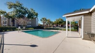 Located at 4700 Walden Cir in Orlando, Florida, this outdoor amenity space serves as the central gathering hub of a residential community. The centerpiece is a generously sized rectangular swimming pool with clear, blue-green water, framed by a wide concrete pool deck ideal for lounging and recreation. A covered pavilion with a pitched roof sits adjacent to the pool, providing shaded relief from the Florida sun and serving as a comfortable outdoor gathering spot. The pool area is fully enclosed by a black wrought iron fence, which includes a gated entry with posted safety signage, including no-diving and no-lifeguard-on-duty notices. Mature palm trees and established shade trees are thoughtfully landscaped throughout the perimeter, adding a lush, tropical character to the environment. Manicured shrubs and green lawn areas soften the hardscape and contribute to the well-maintained appearance of the grounds. The surrounding multi-story residential buildings feature arched windows, balconies, and neutral exterior siding, providing an attractive backdrop to the outdoor space. Under the open Florida sky, this pool area benefits from abundant natural sunlight, creating a bright and inviting atmosphere suited for year-round outdoor enjoyment.