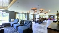This sophisticated single-floor space seamlessly blends comfort and elegance in an open, airy layout designed for both relaxed gatherings and formal occasions. The lounge area is furnished with plush, tufted blue velvet sofas and accent chairs arranged to encourage conversation, while a richly patterned area rug anchors the seating arrangement and adds a touch of warmth. Adjacent to the lounge, a grand formal dining area features a long, dark wood table surrounded by high-backed upholstered chairs, ideal for hosting large groups. A series of large, arched windows line the walls, flooding the interior with abundant natural light and offering picturesque views of lush greenery outdoors. Sheer and patterned drapery in gold tones frame the windows, adding softness and depth to the design. The light-toned tile flooring, laid in a striking hexagonal pattern, transitions beautifully between zones while maintaining visual continuity. Overhead, an impressive array of ornate medallion-style flush-mount light fixtures are distributed across the white ceiling, providing warm, ambient illumination and serving as a distinctive decorative feature. The overall ambiance is refined and inviting, combining classical design elements with a modern sensibility.