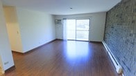 This single-floor space offers a clean, open layout defined by rich, dark hardwood-style flooring that extends throughout the main living area, lending warmth and a polished aesthetic to the interior. The space is bathed in natural light, thanks to a large sliding glass door that serves as both a focal point and a functional access point to a private balcony overlooking outdoor greenery — a detail that brings a sense of openness and connection to the outdoors.

The walls are finished in a crisp, neutral white tone, creating a bright and versatile backdrop that complements a variety of furnishing styles. A wall-mounted air conditioning unit provides climate control, while a baseboard heater along the perimeter ensures comfortable heating year-round. The ceiling is smooth and unadorned, maintaining a clean and minimalist character throughout.

The generous proportions of the main room allow for flexible furniture arrangements, making it well-suited for a variety of living configurations. The overall ambiance is light, airy, and move-in ready, with the combination of natural light, neutral finishes, and durable flooring creating a welcoming and functional environment ideal for everyday living.