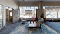 The Kirk Hotel L presents a well-appointed single-floor layout designed to accommodate both social and administrative functions. The expansive main lobby serves as the heart of the space, featuring a prominent front desk framed by distinctive glass block accents and a structural column that anchors the open floor plan. Patterned grey carpet tiles flow consistently throughout, lending a cohesive, polished appearance to the entire level.

A welcoming lounge area near the large street-facing windows offers abundant natural light and views of the surrounding neighborhood. Comfortable seating arrangements, including upholstered chairs and a central coffee table atop a vibrant area rug, create an inviting atmosphere for residents or guests. A separate seating nook features a classic rolled-arm sofa, wall sconces, and a decorative mirror, adding a refined touch.

Additional highlights include a billiards area with a full pool table, a secondary lounge with armchairs and colorful artwork, and a private office equipped with a large desk, wall clock, and ample natural light from street-facing windows. A staircase with ornamental iron railings connects to upper floors. Glass block wall panels and warm wood-framed doors contribute architectural character throughout.
