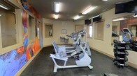 This fitness room offers a practical and well-appointed space designed to support a variety of cardio-focused workouts. The room is outfitted with multiple pieces of cardio equipment, including treadmills, an elliptical trainer, and a stationary recumbent bike, providing users with a range of exercise options. Overhead fluorescent lighting ensures the space is well-lit throughout, while wall-mounted televisions offer entertainment during workouts. A standout visual feature is the bold, colorful mural adorning one wall, adding an energetic and artistic element to the otherwise functional environment. Framed mirrors are positioned along the same wall, helping to amplify the sense of space. Windows on the opposite wall allow for natural light to filter in, contributing to a bright and welcoming atmosphere. The flooring is finished with dark rubber matting, a practical choice suited to heavy equipment and active use. Small utility items such as sanitizing stations are thoughtfully placed for convenience. The overall ambiance is clean, functional, and motivating, making this fitness room a well-considered amenity for its users.