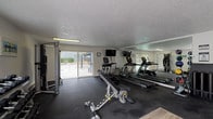 This single-floor fitness center offers a comprehensive workout environment designed to accommodate a wide range of exercise routines. The main workout area features durable rubber flooring throughout the equipment zone, transitioning to light wood-look flooring in the adjoining entry and transitional spaces. A textured ceiling runs throughout, and the room is illuminated by overhead fluorescent lighting supplemented by natural light from multiple windows and a sliding glass door.

The cardio section is anchored by multiple True treadmills and a stair climber, positioned in front of a large wall-to-wall mirror that enhances the sense of space and allows users to monitor their form. A wall-mounted television is installed nearby for entertainment during workouts. Strength training is well-supported by a Hoist functional cable trainer, a multi-position adjustable bench, and a rack of hex dumbbells in varying weights. A medicine ball storage rack holds an assortment of weighted balls for functional training, along with foam rollers for recovery.

A transitional area connects to additional rooms and features a mini-split HVAC unit for climate control, a storage locker, and a TRX stability ball. The sliding glass door opens directly to an outdoor patio and fenced area, providing convenient access to the exterior.