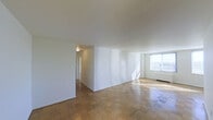 This single-floor space is characterized by its clean, minimalist design and consistent use of classic parquet wood flooring throughout, lending warmth and a timeless quality to each area. The walls are finished in crisp white, contributing to a bright, neutral palette that enhances the sense of openness across the layout.

The main living area is notably spacious, benefiting from a large window that allows abundant natural light to flood the interior, casting sunlight across the parquet floors and illuminating the room's generous proportions. A baseboard heating unit is visible beneath the window, indicating a practical heating solution integrated into the space.

A partial wall divides the main living area from an adjoining hallway or secondary space, maintaining a degree of definition between zones while preserving an open feel. The entry area features a louvered closet door and a standard entry door, providing functional storage directly upon arrival. An electrical panel is mounted near the entry, and standard electrical outlets are positioned throughout.

The ceilings are smooth and uniform in height, reinforcing the clean architectural lines of the space. Overall, the unit presents a straightforward, functional layout with a light-filled, uncluttered ambiance well-suited for a variety of configurations.