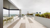 Located at 501 Broadway in Santa Monica, California, this expansive rooftop terrace offers a sophisticated open-air amenity space designed for both relaxation and social gatherings. The terrace is finished with large-format porcelain tiles arranged in a two-tone pattern, lending a clean, modern aesthetic throughout.

Multiple distinct seating zones are thoughtfully distributed across the rooftop. A lounge area centered around a round concrete fire pit provides a cozy gathering spot, surrounded by cushioned chairs ideal for evening use. A separate sectional seating arrangement with green-framed furniture offers additional space for casual socializing, positioned to take full advantage of sweeping views toward the Pacific Ocean and the Santa Monica Pier. A covered pergola with a dark metal frame shelters an outdoor dining table and chairs, providing a shaded setting for alfresco meals alongside a built-in prep counter.

Generously sized raised planters in a muted gray finish are strategically placed across the terrace, filled with a variety of drought-tolerant shrubs, ornamental grasses, and slender trees that add vertical interest and natural greenery. Low-profile lounge chairs are also available for sunbathing or taking in the panoramic skyline and coastal views. The overall design is contemporary and cohesive, reflecting the vibrant yet laid-back character of its Santa Monica surroundings.
