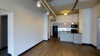 This single-floor space showcases a striking industrial-modern aesthetic, characterized by soaring ceilings with exposed ductwork and raw concrete elements overhead. The open-concept layout seamlessly connects the living area to the kitchen, creating a spacious and versatile environment. Warm hardwood-style flooring runs throughout, grounding the space with a natural contrast to the industrial ceiling details.

The kitchen is a standout feature, equipped with light gray cabinetry that extends to the upper walls, dark granite or stone countertops, and black appliances including a full-size refrigerator, microwave, and range. A kitchen peninsula provides additional prep space and serves as a natural divider between the cooking and living zones. Pendant lighting hangs above the peninsula, offering focused illumination and a contemporary design touch.

The living area is generously proportioned, with clean white walls that allow for flexible furnishing arrangements. Recessed and track lighting supplements the pendant fixtures, ensuring consistent brightness throughout. The overall ambiance is modern and urban, balancing raw industrial elements with clean, polished finishes.