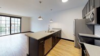 This single-floor, one-bedroom, one-bathroom unit showcases a contemporary design with consistent light wood-tone flooring and bold dark baseboards throughout. The open-concept kitchen serves as a central highlight, featuring dark cabinetry, quartz countertops, a subway tile backsplash, and a full suite of stainless steel appliances including a refrigerator, range, microwave, and dishwasher. A peninsula island with pendant lighting overhead provides additional prep space and a natural division between the kitchen and living area. The living space is bright and airy, benefiting from large, multi-pane sliding glass doors that flood the interior with natural light and offer views of the surrounding neighborhood. The bedroom continues the clean aesthetic with its own generously sized window and direct access to a spacious walk-in closet outfitted with multi-level wire shelving and hanging rods for organized storage. The bathroom presents a refined finish with a glass-enclosed walk-in shower clad in large-format dark tile, complemented by a grey vanity with dark countertop and rain shower fixtures, completing the unit's modern, cohesive design.