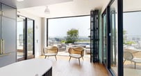 This elegantly designed single-floor space occupies an elevated position within a high-rise building, offering sweeping panoramic city views through floor-to-ceiling windows that flood the interior with natural light. Wide-plank hardwood flooring runs throughout, anchoring a cohesive and refined aesthetic.

At the heart of the space is a striking elongated bar featuring a sculpted blue fluted base and a crisp white surface, surrounded by upholstered metal barstools. Behind it, a full kitchen wall finished in steel-blue cabinetry frames a dramatic book-matched marble slab backsplash, illuminated by warm accent lighting.

A glass-walled conference room with sliding dark-framed panels provides a dedicated meeting area while maintaining visual connectivity to the open floor plan. Sheer drapery allows for privacy when needed. Adjacent to this, a comfortable lounge area offers relaxed seating, a large wall-mounted display, and bold accent chairs in deep red.

The space extends outdoors to a generously sized terrace furnished with sectional seating, lounge chairs, a central fire pit, and lush planted greenery — all set against an expansive urban skyline backdrop, making it ideal for both professional gatherings and informal entertaining.