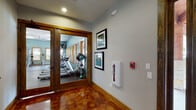 This single-floor fitness center is thoughtfully designed to support a comprehensive workout experience. The entry corridor sets an inviting tone with richly polished concrete floors in warm amber tones, light gray walls adorned with framed artwork, and warm wood baseboard trim. A set of wood-framed glass double doors provides a clear view into the main exercise area, building anticipation before entry.

Inside, the spacious workout floor is finished with durable speckled rubber flooring and features teal-painted walls that lend a calm, energizing atmosphere. Wood-trimmed windows line the walls, flooding the space with natural light and offering views of the surrounding outdoor environment. Ceiling fans and fluorescent lighting ensure the space remains well-ventilated and brightly lit throughout.

The cardio zone is generously stocked with multiple treadmills, elliptical trainers, and stationary bikes, arranged to take full advantage of the window views. A large wall mirror on one side visually expands the space and is useful for monitoring form. The strength training area includes a cable machine, squat rack, weight bench, dumbbells, kettlebells, and medicine balls, offering a full range of resistance training options. Wall-mounted televisions add convenience for entertainment during workouts. Safety features include a mounted fire extinguisher and alarm system near the entrance.