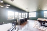 Located at 501 North Christopher Columbus Boulevard in Philadelphia, this well-appointed game and lounge room offers a sophisticated recreational experience with striking design details throughout. The space is defined by its rich deep-green and sage-toned walls, complemented by elegant wainscoting paneling that adds a refined architectural character. Recessed lighting and a modern multi-arm chandelier illuminate the room, while large windows along multiple walls flood the interior with natural light and frame sweeping panoramic views of the surrounding waterfront and Philadelphia skyline. The tile flooring, set in a diagonal pattern, anchors the space with a classic yet contemporary feel. A full-size pool table positioned atop a wave-patterned area rug serves as the room's central feature, making it ideal for entertainment. A wall-mounted flat-screen television with built-in cabinetry provides a dedicated media area, while a long bar-height counter with bar stools offers a comfortable spot for socializing. A round game table with wicker seating adds further versatility. Glass doors lead to an outdoor terrace, seamlessly extending the lounge experience to the outside. The overall ambiance is upscale and inviting, blending modern amenities with refined interior design.