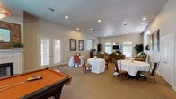 This well-appointed single-floor gathering space is designed to accommodate a variety of social activities in a warm, inviting environment. The layout flows seamlessly across distinct functional zones, each thoughtfully furnished and bathed in natural light from numerous windows fitted with blinds.

At the heart of the space is a generously sized game room anchored by a full-sized pool table and a classic white-mantel fireplace, creating a focal point that is both functional and aesthetically pleasing. A ceiling fan overhead and a curved bank of windows enhance the airy feel of this area.

Adjacent to the game room, a comfortable lounge area features plush, upholstered seating, framed artwork, and a large flat-screen television. Arched windows with transom lights draw in abundant natural light and provide views to the outdoors. French doors offer direct access to the exterior, further connecting the interior to the outside environment.

Round dining tables draped in white linens are arranged to accommodate group gatherings, complemented by classic wood chairs. A pass-through window connects this area to a compact service kitchen equipped with white cabinetry, a range, and counter seating for two — ideal for catering or light meal service.

Throughout, neutral carpet flooring, recessed lighting, and warm beige walls create a cohesive and welcoming ambiance suited for community events, private gatherings, or recreational use.