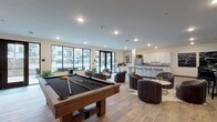 Located at 40 Hutchinson Ave in Columbus, Ohio, this single-floor community amenity space is thoughtfully designed to offer residents a modern, upscale environment for relaxation and socializing. The open-concept layout flows seamlessly between distinct zones, each offering a unique purpose while maintaining a cohesive contemporary aesthetic throughout.

The game and social lounge area features a full-size pool table as its centerpiece, surrounded by plush swivel lounge chairs and accented by large windows and French doors that flood the space with natural light and provide views of the outdoor courtyard. Adjacent to this is a fully appointed kitchen with crisp white cabinetry, granite countertops, a central island with bar seating, stainless steel appliances, and ample prep space — ideal for entertaining.

A separate lounge area anchored by a textured stone accent wall houses a linear gas fireplace flanked by wall-mounted flat-screen TVs. Deep leather sofas, accent chairs in rich emerald green, and a statement chandelier create a warm, sophisticated atmosphere perfect for gathering.

Glass-partitioned spaces near the entry provide semi-private seating with high-top tables and bistro chairs, framed by bold abstract artwork. The tile and wood-look flooring throughout ties each zone together, delivering a polished, well-appointed clubhouse experience.