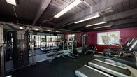 This fitness center offers a comprehensive workout environment designed to support a full range of exercise routines. The space is defined by its bold, two-tone color scheme — deep crimson and teal accent walls contrast against a dark, beam-exposed ceiling, giving the gym a dynamic and energizing atmosphere. Durable rubber flooring spans the entire space, providing a safe and practical surface for all types of training.

Along the front wall, large floor-to-ceiling sliding glass doors and windows open to views of an outdoor pool and patio area, flooding the gym with natural light and creating a seamless connection to the exterior. Overhead, industrial-style fluorescent lighting ensures the space is well-lit throughout.

Cardio equipment, including multiple treadmills and an elliptical machine, is positioned to take advantage of the poolside views. The strength training section features a multi-functional cable machine with a full instructional guide, a weight bench, a stability ball, and a selection of medicine balls and free weights. Full-length mirrors line one wall, enhancing the sense of space and allowing users to monitor their form during workouts. A utility table is also available for convenience. The gym is a well-rounded facility suited for both cardio and strength-focused training sessions.