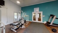 Located at 151 Century Dr in Greenville, South Carolina, this fitness center offers a well-equipped, single-floor exercise facility designed to support a range of workout routines. The space is bright and welcoming, with white walls accented by a bold teal feature wall that adds a lively, energizing atmosphere. Large windows and glass-paneled double doors allow natural light to fill the room, providing pleasant views of the surrounding outdoor greenery.

The fitness center is furnished with a comprehensive selection of equipment, including a treadmill, elliptical machine, and multiple cable and weight machines suitable for both strength training and functional fitness. A wall-mounted television is positioned for convenient viewing during workouts. A large mirror along one wall enhances the sense of space and allows users to monitor their form. Dumbbells and a stability ball round out the equipment offerings, catering to varied fitness preferences.

Durable carpet flooring with area rugs defines workout zones throughout the room. The facility also features a hand sanitizer dispenser, posted fitness center rules, and recessed lighting overhead, ensuring a clean and organized environment. The double-door entry with decorative glass panels provides an inviting access point, with outdoor views hinting at proximity to additional community amenities.