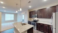 This single-story space showcases a well-appointed kitchen as its centerpiece, designed with both functionality and aesthetics in mind. The kitchen features rich, dark espresso-toned cabinetry that extends from floor to ceiling, providing generous storage throughout. Light granite countertops offer a striking contrast against the dark cabinets, running along the perimeter and continuing onto a central island that includes a stainless steel sink and serves as an ideal space for meal preparation or casual dining. Pendant lighting hangs above the island, adding a refined touch to the workspace. Stainless steel appliances, including a built-in microwave, electric range, and oven, complete the kitchen's modern character. The open layout connects the kitchen seamlessly to the living area beyond, which benefits from natural light streaming in through multiple windows. Neutral-toned walls and light wood-style flooring flow throughout, creating a cohesive and airy atmosphere. The overall design balances practicality with contemporary style, making the space feel both inviting and functional.