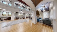 Located at 13125 Watertown Plank Road in Elm Grove, Wisconsin, this remarkable single-floor space is a masterfully repurposed historic building that seamlessly blends classical ecclesiastical architecture with contemporary design. The grandeur of the space is immediately apparent upon entry, with soaring vaulted and domed ceilings accented by rich dark wood detailing and sweeping arches that draw the eye upward throughout.

Ornate white columns with intricately carved capitals define distinct functional zones across the expansive open floor plan, all unified by warm hardwood flooring. Multiple seating areas are thoughtfully arranged, including lounge groupings with leather sofas and armchairs, as well as a formal dining area centered around a solid wood table. Decorative area rugs anchor each arrangement, adding texture and warmth.

Original stained glass windows line the walls, filling the interior with filtered, colorful natural light and serving as striking focal points throughout. Arched niches display classical artwork, preserving the space's historic character.

The fully equipped kitchen features a large island with an elegant marble-look countertop, a gas cooktop, and modern cabinetry, harmoniously integrated within the ornate surroundings. The overall ambiance is one of refined grandeur — an inspiring blend of historic preservation and thoughtful modern functionality.