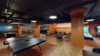 This single-floor entertainment space is defined by a cohesive design language of deep terracotta walls, black exposed ceilings, and warm tile flooring that flows throughout its interconnected zones. The open main area anchors the space with a ping-pong table at its center, surrounded by eclectic seating including lounge chairs and counter-height tables with teal barstools, creating a casual, social atmosphere. A dedicated bowling area features two lanes accompanied by black leather sofas, rich burgundy walls, and dramatic curtains for an intimate, venue-like feel. The full wet bar serves as a sophisticated focal point, boasting a sweeping dark granite countertop, navy velvet barstools with gold-toned frames, textured mosaic wall tiles, and mirrored backsplash panels that amplify the room's depth. A curved lounge nook provides a more intimate retreat, with built-in banquette seating, pendant lighting, a mounted flat-screen television, and arched walls that soften the overall industrial aesthetic. Throughout the space, rounded terracotta columns, a wet kitchenette, a backgammon corner, and carefully placed accent lighting contribute to a layered, upscale entertainment environment.