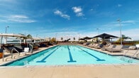 Located at 1000 Elements Way in Irvine, California, this outdoor amenity space presents a resort-inspired pool deck designed for both recreation and relaxation. The centerpiece is a generously sized lap pool with crystal-clear turquoise water and clearly marked swim lanes, ideal for both fitness and leisure. The pool deck is finished with large-format, neutral-toned pavers that lend a clean, contemporary aesthetic throughout the space. Surrounding the pool, an extensive lounge area is outfitted with cushioned chaise loungers and oversized shade umbrellas, providing comfortable spots to unwind under the Southern California sun. Lush potted palms and planters with colorful foliage add a touch of greenery and help define the space. The open-air design allows for unobstructed views of the surrounding Irvine skyline and the expansive blue sky above, enhancing the bright and airy atmosphere. Additional seating arrangements are thoughtfully distributed around the perimeter, creating social gathering areas for residents or guests. The overall ambiance is upscale and inviting, combining the functionality of a lap pool with the relaxed luxury of a resort-style retreat in the heart of Irvine's urban landscape.