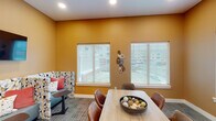 This single-floor space is designed to serve as a versatile gathering and collaborative area, combining a central meeting zone with comfortable lounge seating. The room is immediately distinguished by its bold, warm golden-yellow walls, which create an energetic and inviting atmosphere throughout.

The central focus of the space is a light wood dining table surrounded by cognac leather chairs, ideal for group meetings or casual gatherings. A decorative bowl centerpiece adds a refined touch to the table's surface. Multiple white-trimmed windows fitted with horizontal blinds line the walls, allowing ample natural light to filter in while offering views of the surrounding exterior.

A standout feature is the built-in booth seating area, composed of three semi-private cubicle-style benches upholstered in teal with patterned geometric dividers. Each booth is equipped with a small side table, and a wall-mounted flat-screen TV is positioned above for shared viewing. Textured carpet flooring runs throughout, adding warmth underfoot.

A wide doorway with sliding or swinging glass-paneled doors connects this room to an adjacent lounge and common area, enhancing the overall flow and functionality of the space.