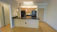 This single-floor space showcases a thoughtfully designed open-concept layout, with the kitchen serving as a central highlight. The kitchen features a large island with a dark granite countertop that provides ample workspace and doubles as a casual dining or gathering area. A stainless steel sink is integrated into the island, while three sleek pendant lights hang overhead, adding both functional illumination and a modern aesthetic touch. An additional fluorescent ceiling fixture ensures the space remains well-lit throughout.

The cabinetry is finished in a warm light maple tone, offering generous storage across both upper and lower configurations. Black appliances, including a refrigerator, electric range, and over-the-range microwave, provide a contemporary contrast against the lighter cabinetry and walls. Dark countertops along the back wall echo the island surface, creating visual cohesion throughout the kitchen.

Light hardwood flooring extends throughout the visible living areas, lending warmth and continuity to the space. The neutral gray walls maintain a clean, understated backdrop. A wood-tone entry door is visible in the background, consistent with the overall modern design palette of the unit.