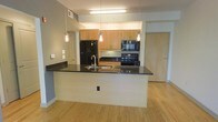 This single-floor space showcases a thoughtfully designed open-concept layout, with the kitchen serving as a central highlight. The kitchen features a large island with a dark granite countertop that provides ample workspace and doubles as a casual dining or gathering area. A stainless steel sink is integrated into the island, while three sleek pendant lights hang overhead, adding both functional illumination and a modern aesthetic touch. An additional fluorescent ceiling fixture ensures the space remains well-lit throughout.

The cabinetry is finished in a warm light maple tone, offering generous storage across both upper and lower configurations. Black appliances, including a refrigerator, electric range, and over-the-range microwave, provide a contemporary contrast against the lighter cabinetry and walls. Dark countertops along the back wall echo the island surface, creating visual cohesion throughout the kitchen.

Light hardwood flooring extends throughout the visible living areas, lending warmth and continuity to the space. The neutral gray walls maintain a clean, understated backdrop. A wood-tone entry door is visible in the background, consistent with the overall modern design palette of the unit.