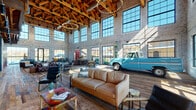 This expansive single-floor space masterfully blends industrial heritage with modern design. The defining architectural feature is the dramatic vaulted ceiling, where exposed heavy timber trusses span the full width of the room, complemented by warm pendant and chandelier lighting that adds an inviting glow to the soaring interior. Whitewashed brick walls line the perimeter, reinforcing the building's industrial character while lending a refined brightness to the space.

Floor-to-ceiling steel-framed windows run along multiple walls, flooding the interior with abundant natural light and providing open views of the surrounding neighborhood. Reclaimed hardwood flooring with rich variation in tone and grain ties together the various zones throughout the space.

The main hall is furnished with multiple distinct lounge areas, including tufted leather Chesterfield sectionals, modern leather sofas, and eclectic seating arrangements, offering both intimate and open gathering spaces. A standout feature is the display of a restored vintage turquoise pickup truck, accompanied by a classic green gas pump, serving as a bold centerpiece. A traditional red British telephone booth adds further character.

A dedicated coffee bar area features a tiled feature wall, sleek counter seating, and full service equipment. Glazed partition walls separate additional lounge and meeting areas, maintaining visual connectivity across the entire space while allowing for functional flexibility.