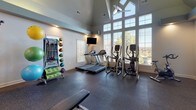Located at 331 Players Club Dr in Castle Rock, Colorado, this single-level fitness center is designed to support a comprehensive workout experience. The space is defined by its dramatically high ceilings and expansive floor-to-ceiling windows that flood the interior with natural light while offering scenic views of the surrounding Colorado landscape. A ceiling fan adds to the airy, open atmosphere of the main exercise area.

The flooring throughout is durable rubber — ideal for both cardio and strength training activities. The cardio zone is well-equipped with multiple treadmills and elliptical machines positioned in front of the large windows, complemented by a wall-mounted flat-screen television. Full-length mirrors line one wall, enhancing the sense of space and allowing for form monitoring during workouts.

The strength training area features a tiered dumbbell rack with a selection of free weights, adjustable weight benches, a multi-station cable machine, and a dedicated rack of stability balls and resistance accessories. Kettlebells and additional functional fitness tools are also available.

Recessed lighting provides consistent illumination throughout, while posted fitness rules and safety signage contribute to an organized, professional environment. An arched doorway connects the main fitness floor to a hallway leading to the entrance, ensuring easy accessibility throughout the facility.