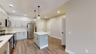 Located at 898 Buffalo Rd in Gates Center, NY, this thoughtfully designed single-floor unit offers a clean, modern aesthetic with consistent finishes throughout. The open-concept kitchen serves as the heart of the space, showcasing gray shaker-style cabinetry, bright white countertops, and a full suite of stainless steel appliances including a refrigerator, range, microwave, and dishwasher. A center island with pendant lighting adds both functionality and visual appeal, while recessed lighting illuminates the space efficiently. Durable wood-look plank flooring runs seamlessly throughout the entire unit, tying each area together with a cohesive, contemporary feel. The living area benefits from sliding glass doors that open to an outdoor view, allowing natural light to fill the room. Additional rooms feature windows with blinds, providing a bright and comfortable atmosphere. The bathroom is well-appointed with a gray vanity, white countertop, matte black fixtures, a large mirror with stylish vanity lighting, and a walk-in shower. Neutral warm-toned walls throughout the unit create a calm and inviting ambiance, offering a versatile backdrop for any style of furnishing.