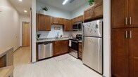 Located at 1401 Forensic Science Dr in Huntington, WV, this single-floor unit offers a well-organized layout that efficiently combines living, sleeping, and cooking spaces. The kitchen is a standout area, outfitted with warm honey-toned shaker-style cabinetry extending to the upper walls for ample storage. Light granite countertops provide generous workspace and complement the full suite of stainless steel appliances, including a refrigerator, electric range, built-in microwave, and dishwasher. Light wood-look flooring runs through the kitchen, contributing to a clean, contemporary feel.

The bedroom is carpeted and comfortably proportioned, accommodating a full-size bed with under-bed storage drawers alongside a wardrobe unit, seating area, and a desk. A window allows natural light to filter in, brightening the neutral white walls and creating a welcoming atmosphere.

The bathroom maintains the same design consistency found throughout the unit, featuring tile flooring, a granite-topped vanity with matching cabinetry, a shower-tub combination, and wall-mounted shelving for additional storage. Overall, the space presents a cohesive, functional design suited for everyday living.