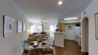 This single-floor space is designed around an open-concept layout that seamlessly connects the dining area, living room, and kitchen into one cohesive, flowing environment. The flooring throughout the main living areas consists of warm wood-style planks, providing a grounded, welcoming aesthetic that contrasts with the light, neutral-toned walls and white ceilings.

The dining area is positioned near the entrance and accommodates a glass-top round table with ornate chairs, making it a functional yet decorative focal point. Beyond it, the living room is generously sized with a ceiling fan providing air circulation, a plush area rug anchoring the seating arrangement, and multiple windows dressed with sheer curtains that allow ample natural light to fill the space.

The kitchen is partially separated by a white peninsula counter that serves as both a workspace and a visual divider. Light wood cabinetry lines the kitchen walls, and a white refrigerator is visible alongside under-cabinet fluorescent lighting. A decorative "Eat" wall sign adds a personalized touch near the kitchen entrance. The overall ambiance is bright, casual, and well-organized.